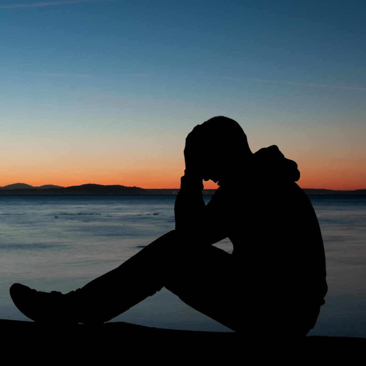 a discouraged man sites at the shore during sunset with his head in his hands